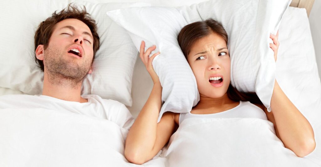 man and woman in bed sleeping. man is snoring loudly. woman covering her ears wishing she has custom earplugs for sleeping